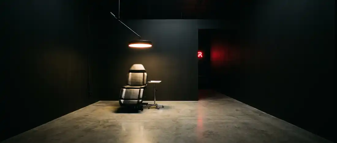 A single tattoo chair lit by one overhead lamp in a dark, near-empty studio.